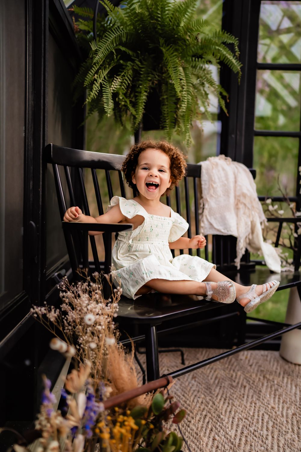 Child in white dress sits on black rocking chair by window with fern plant and dried flowers.