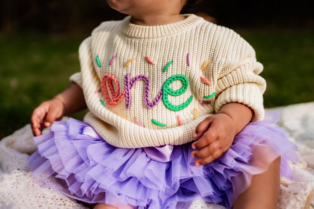 A cream colored knit sweater with colorful text embroidery paired with a purple ruffled tulle skirt.