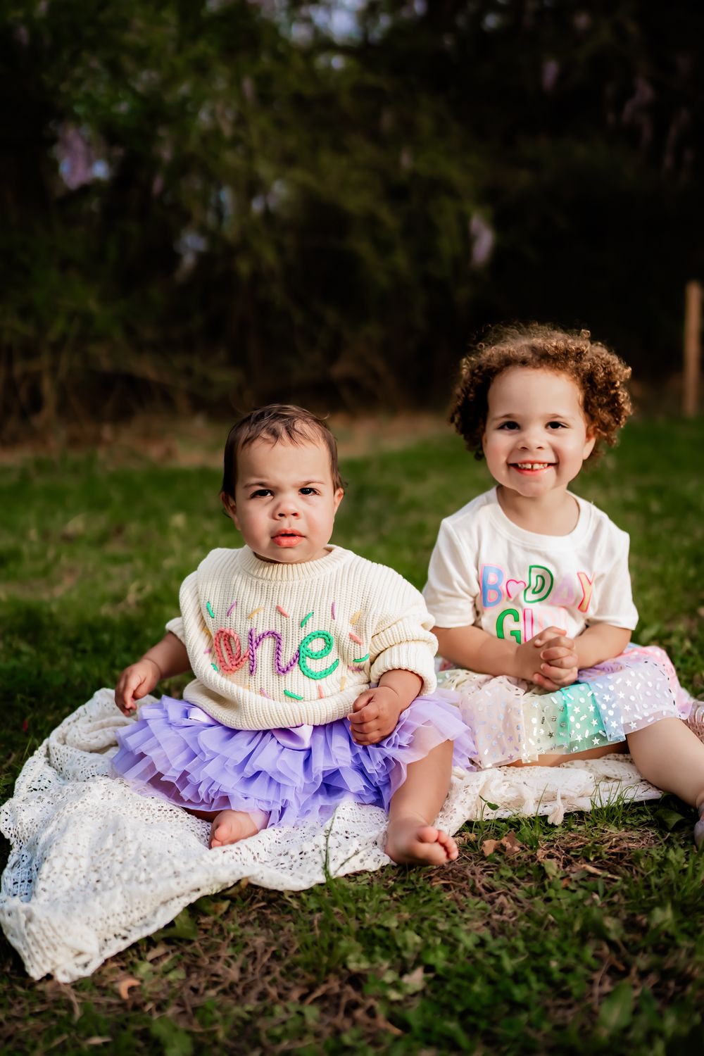 Two smiling children in tulle skirts and custom birthday shirts sit outside on a blanket in a garden setting.