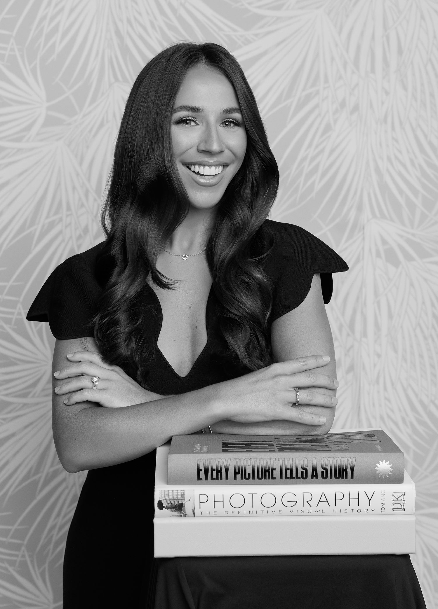 Black and white portrait of a woman in dark dress smiling while posing with photography books against floral wallpaper.