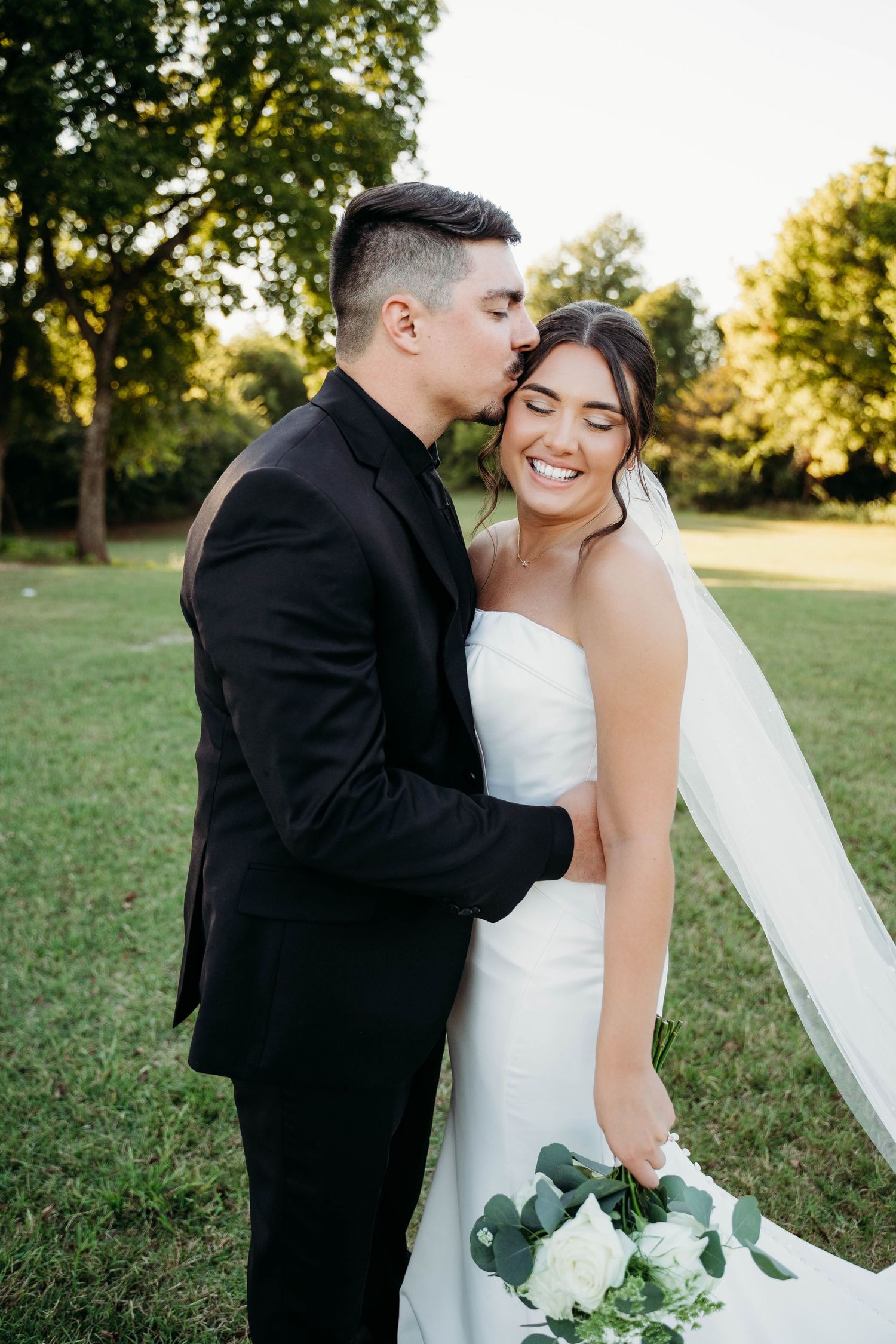 A romantic wedding moment captured outdoors with a bridal bouquet of white roses and greenery on a sunny day.