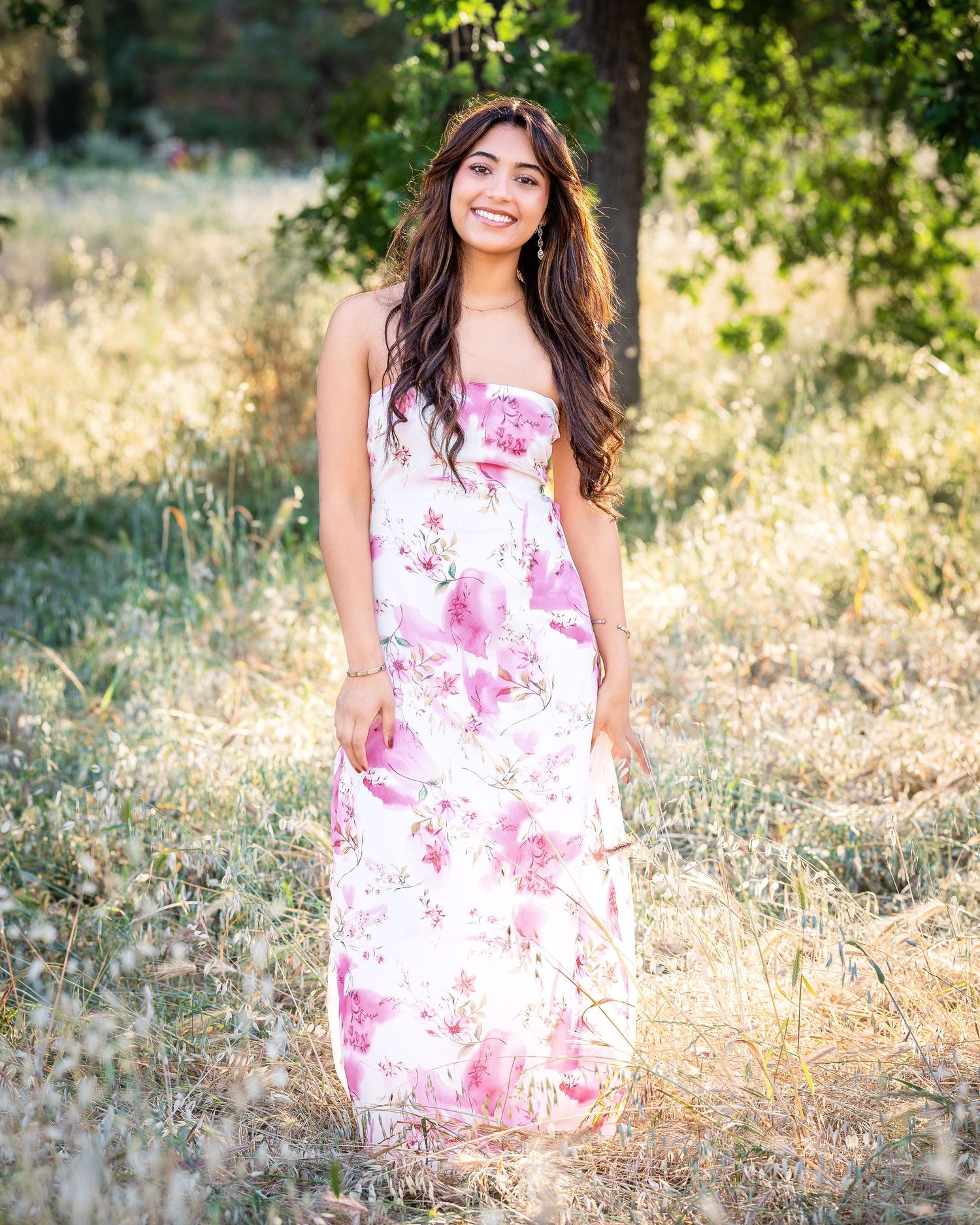 Person in a flowing white and pink floral maxi dress poses outdoors in a sunlit field with golden grass and trees.