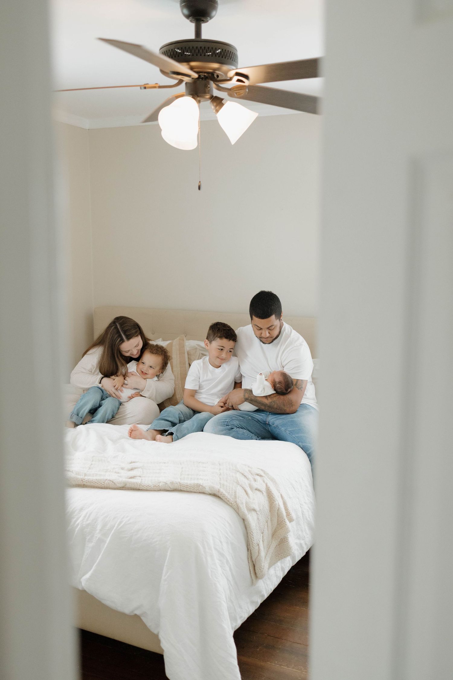 Family relaxing together on a white bed under a ceiling fan in a cozy bedroom.