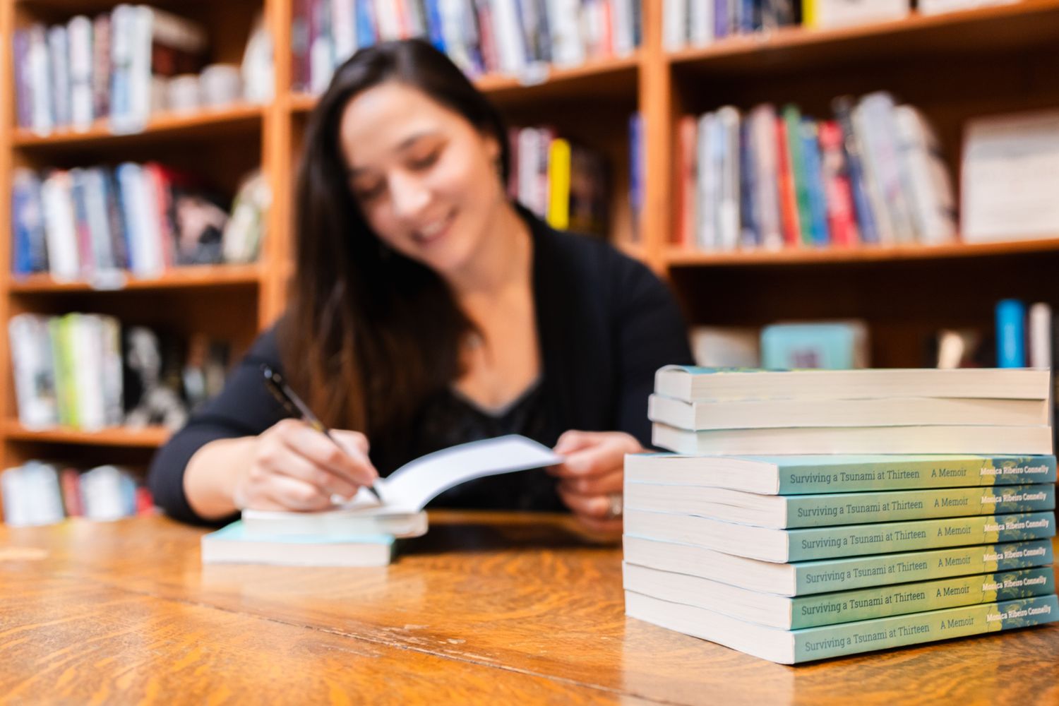 Someone studies at a wooden table in a library with a stack of identical textbooks visible in the foreground.