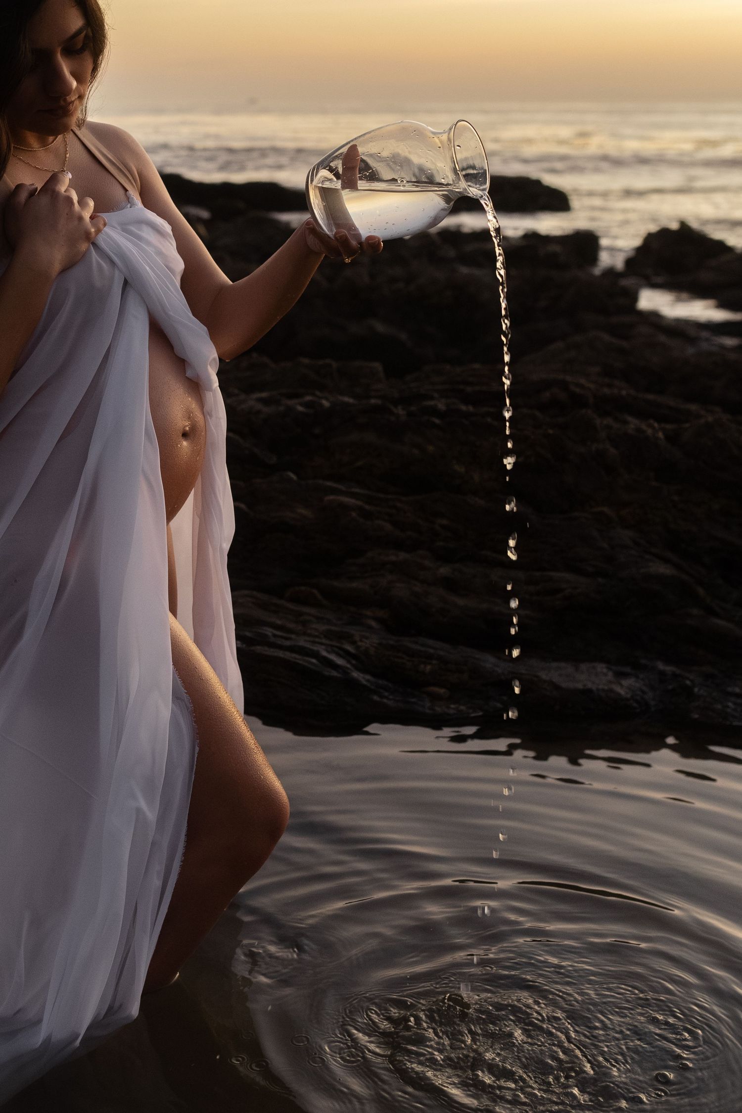 Artistic water droplet photography at sunset with flowing white fabric and rocky coastline.