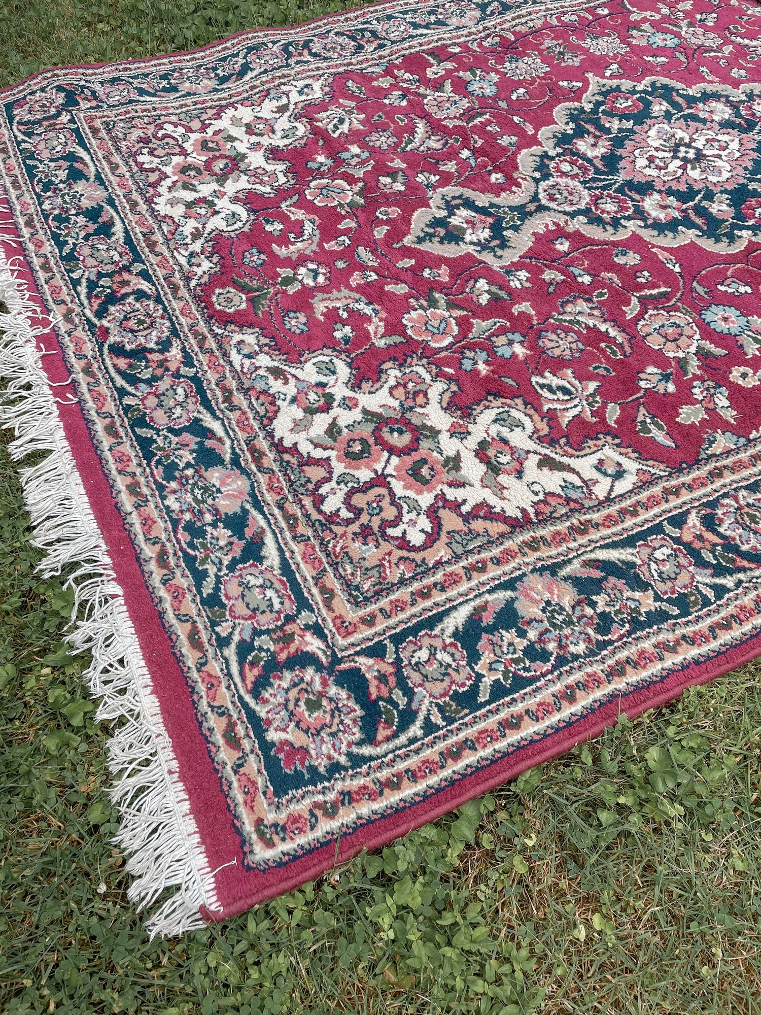 A traditional Persian-style area rug with red and navy floral design features fringe edges and ornate border details on grass.