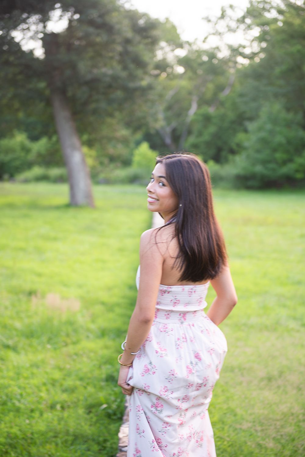 A person wearing a white floral dress poses outdoors in a grassy field with trees in the background.