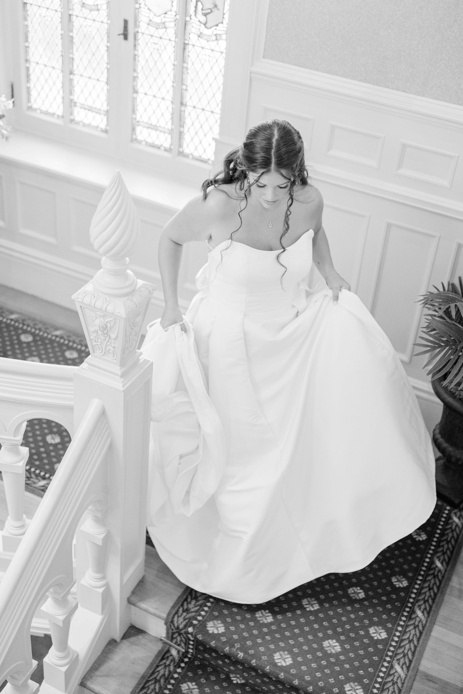 A bride in a white wedding dress sits on a patterned chair in a bright, sunlit room.