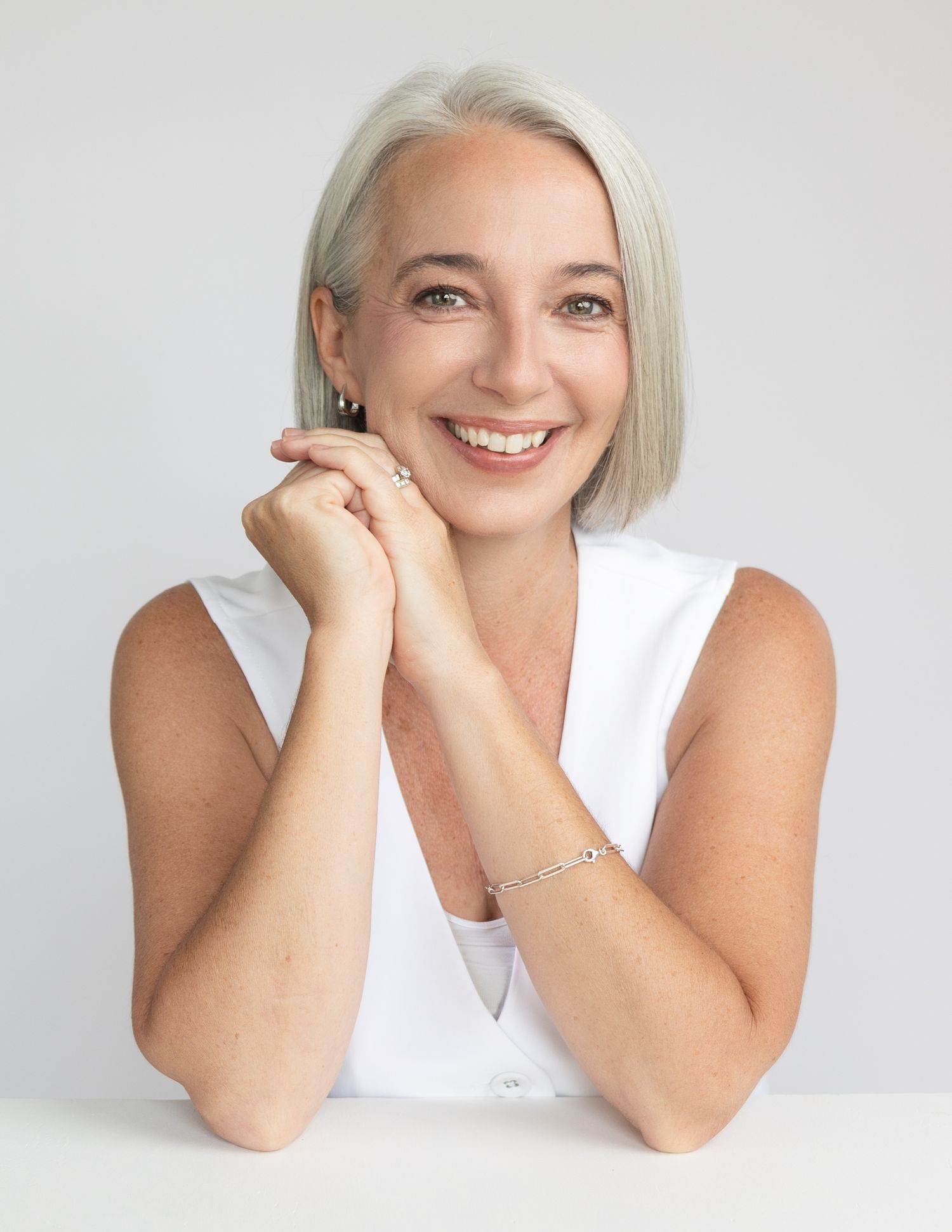 woman with silver hair and green eyes in white button up vest during a branding headshots session leaning on elbows and smiling big.