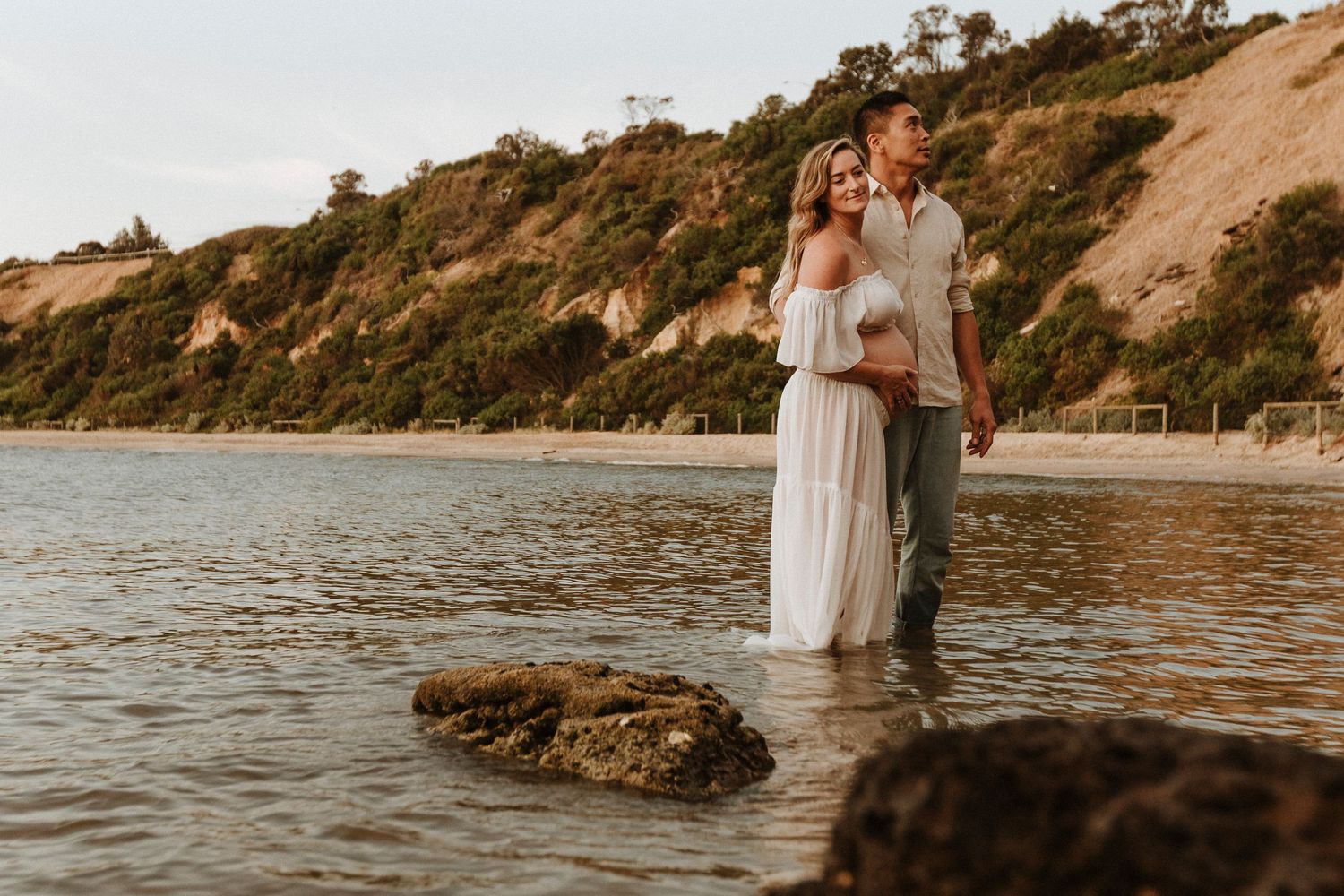 A romantic lifestyle image of a maternity session with two people embracing while standing in shallow ocean water at sunset near cliffs.