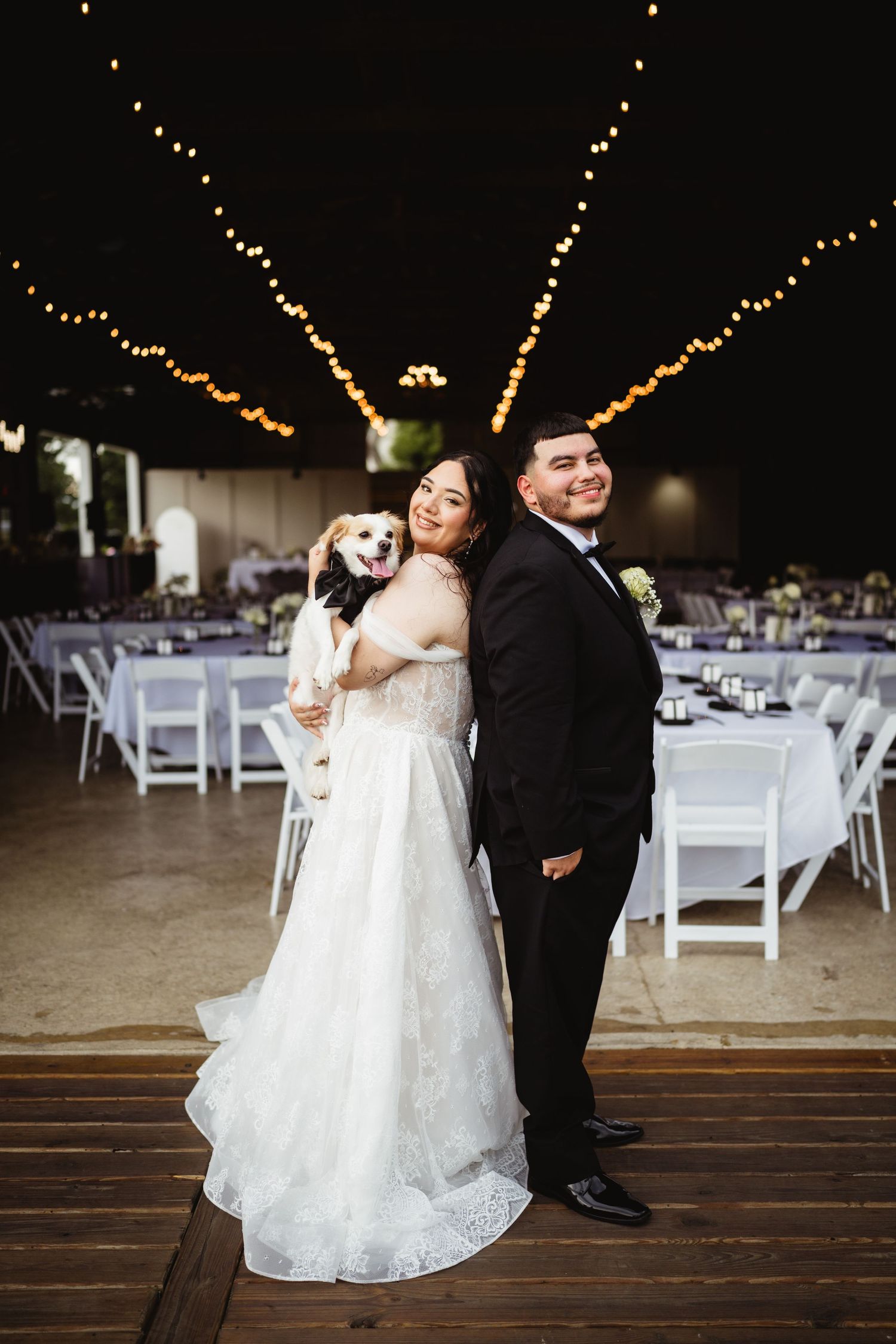 A wedding couple poses with their happy dog at their reception venue decorated with string lights and white chairs.