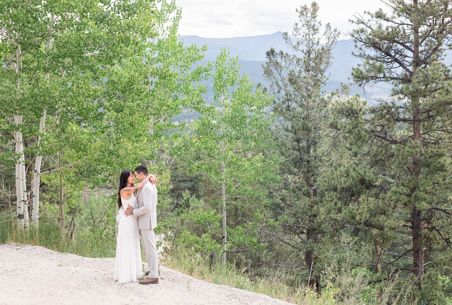 A couple embraces on a mountain path surrounded by evergreen trees and aspens with mountains in the distance.