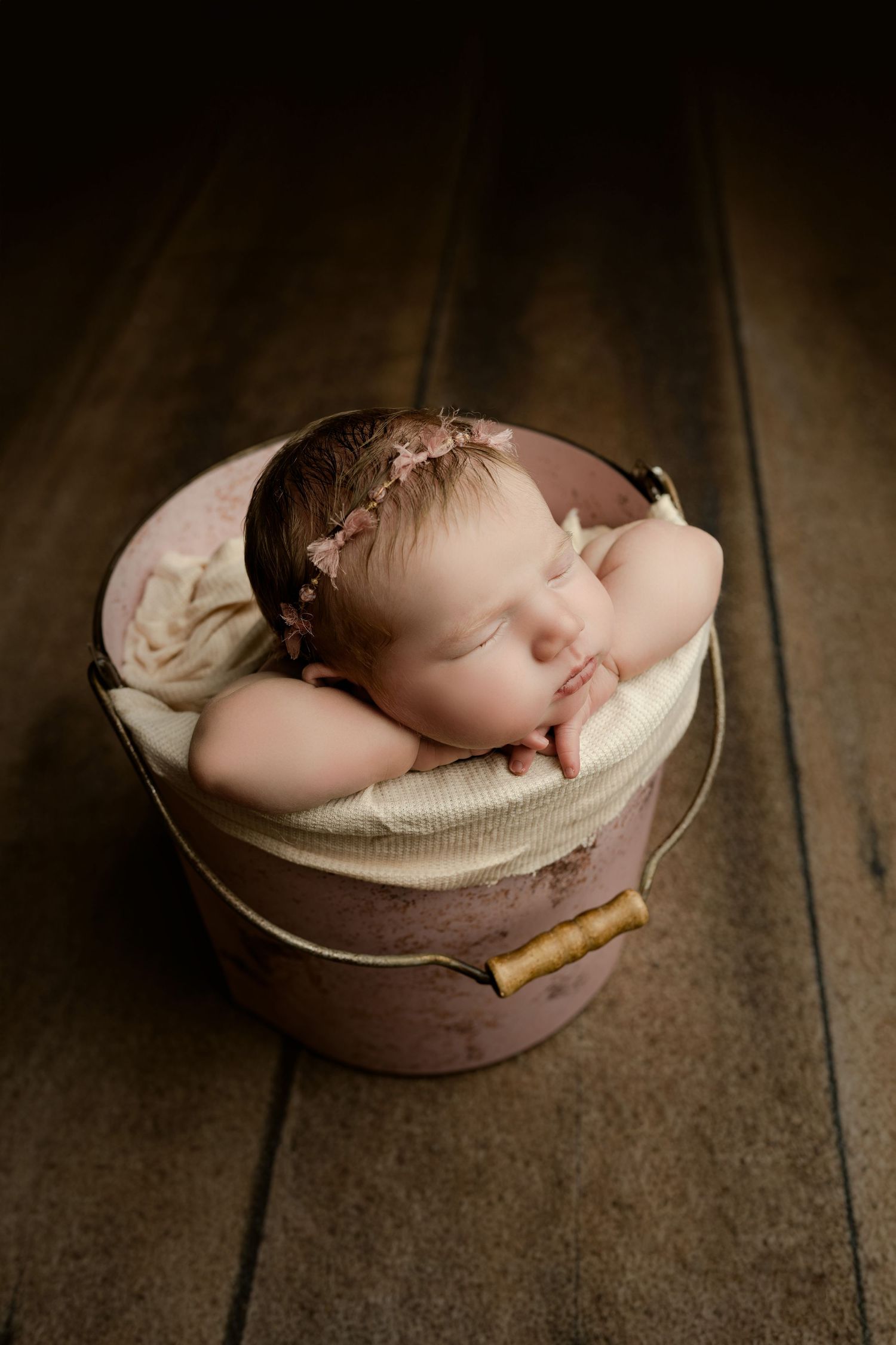 Sleeping newborn baby girl in a pink bucket with a floral headband on a dark wooden floor.