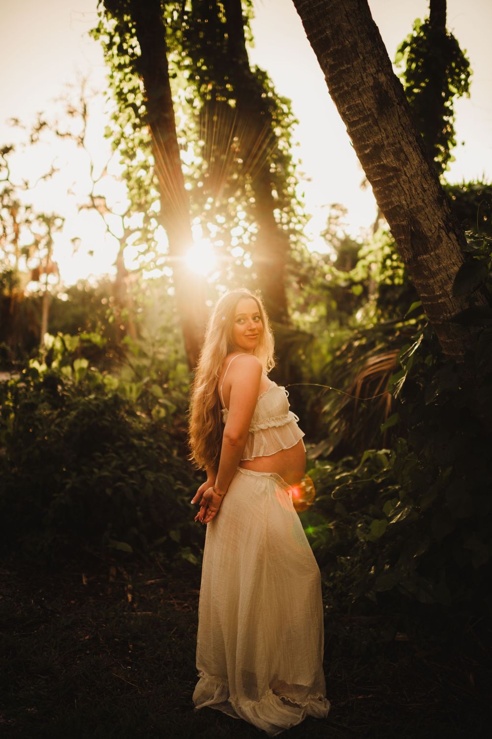 A dreamy backlit portrait of someone in a white dress standing among trees at golden hour sunset. Sarasota best family photographer 2026