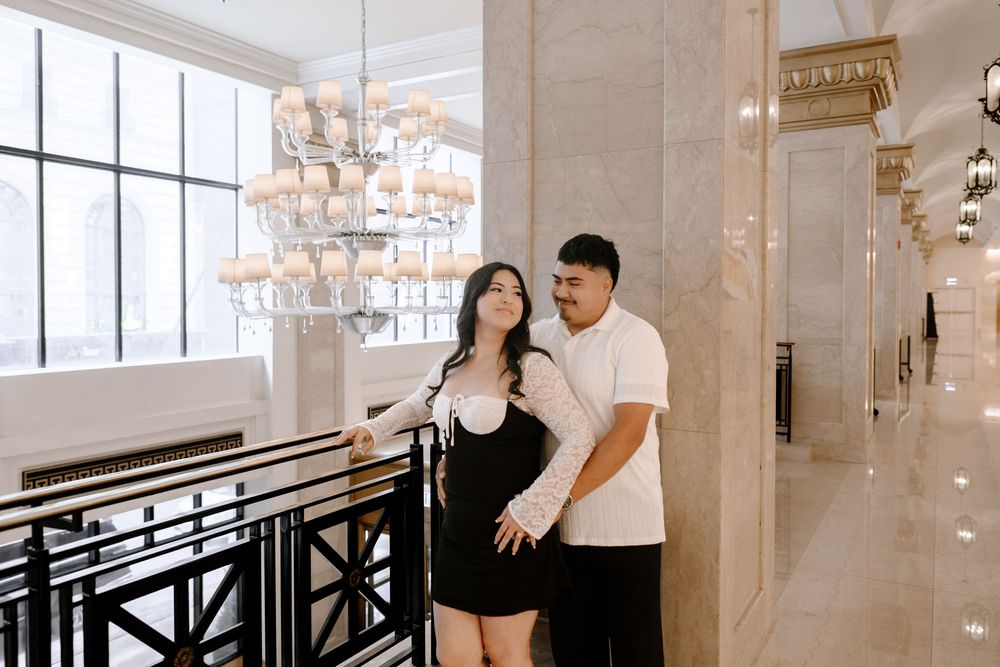 A loving couple poses together on a grand staircase with elegant interior decor and ornate lighting fixtures.