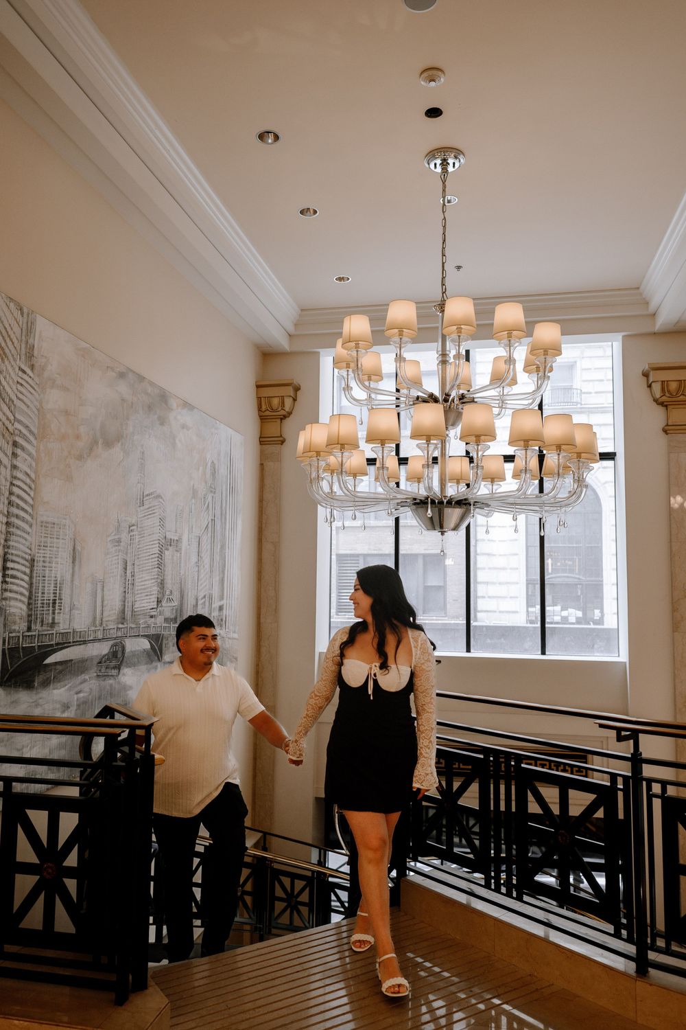 Two people sit on a wooden bench in an elegant room with white walls and hanging chandeliers.