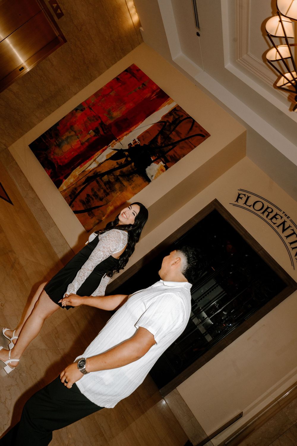 People dance together at a party or celebration with red wall art and overhead lighting visible in the background.