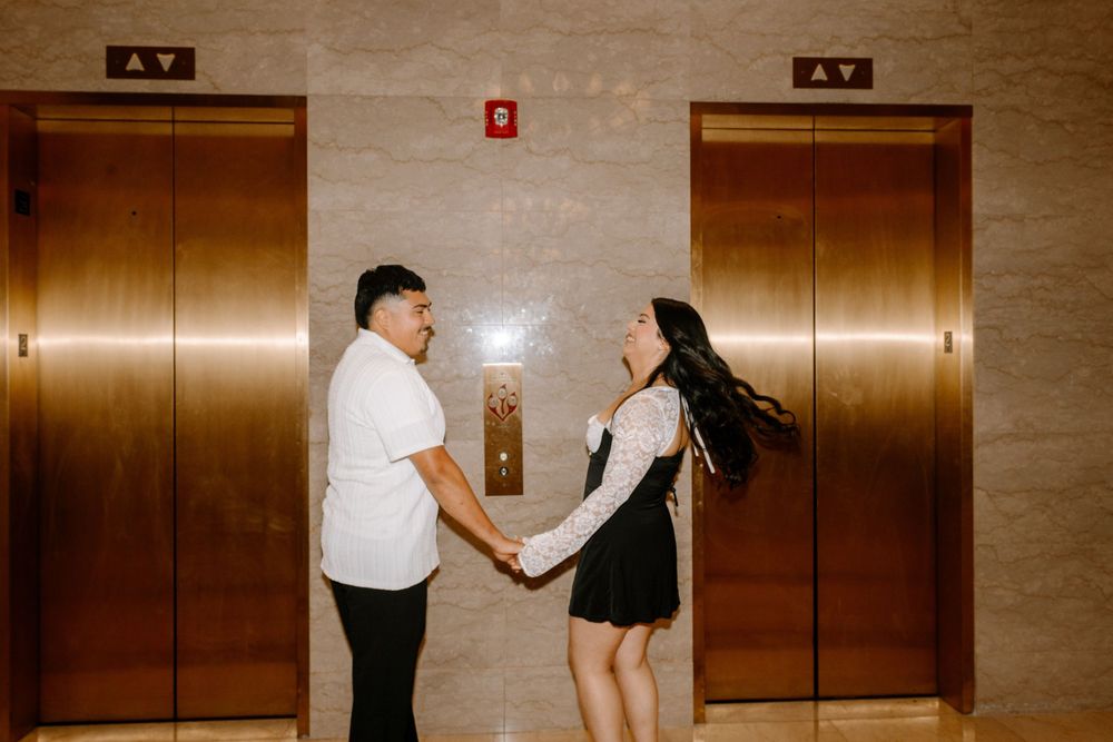 A couple holds hands while standing between two copper-colored elevator doors in a marble hallway.