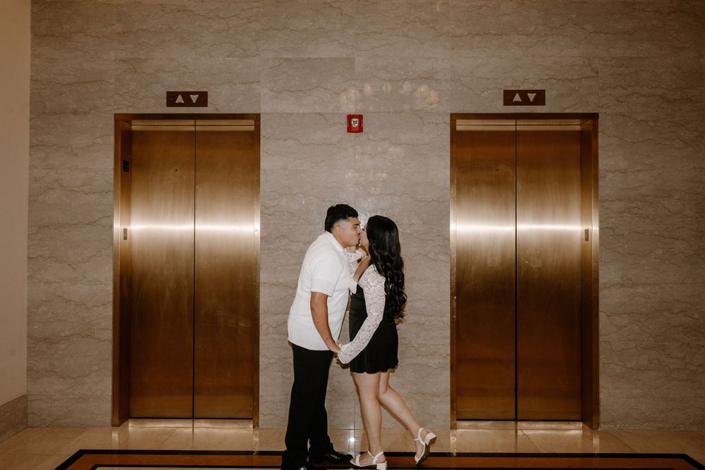 A couple in contrasting black and white outfits embraces between two brass elevator doors in a marble hallway.