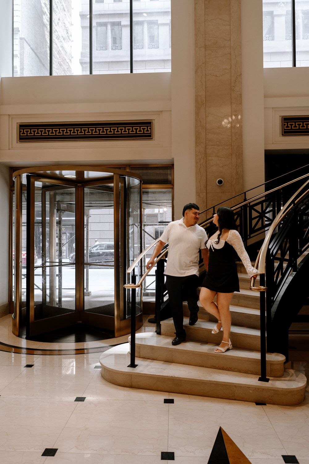 An elegant ballroom with marble columns and black metal railings features ornate chandeliers and classical architectural details.