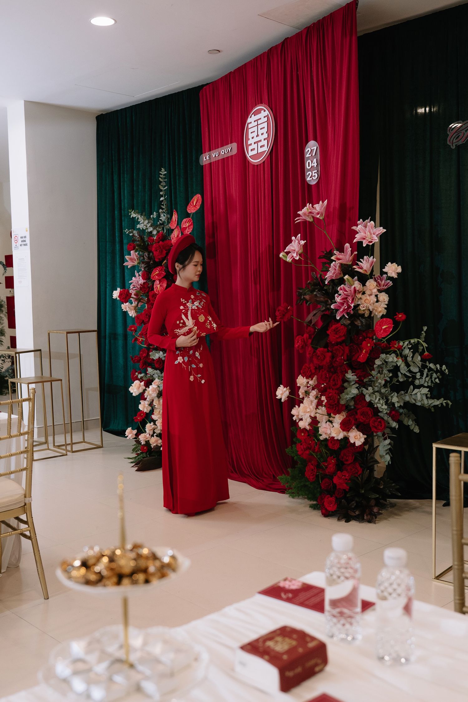 Woman in red ao dai stands before a traditional Vietnamese wedding backdrop with red curtains and floral arrangements.