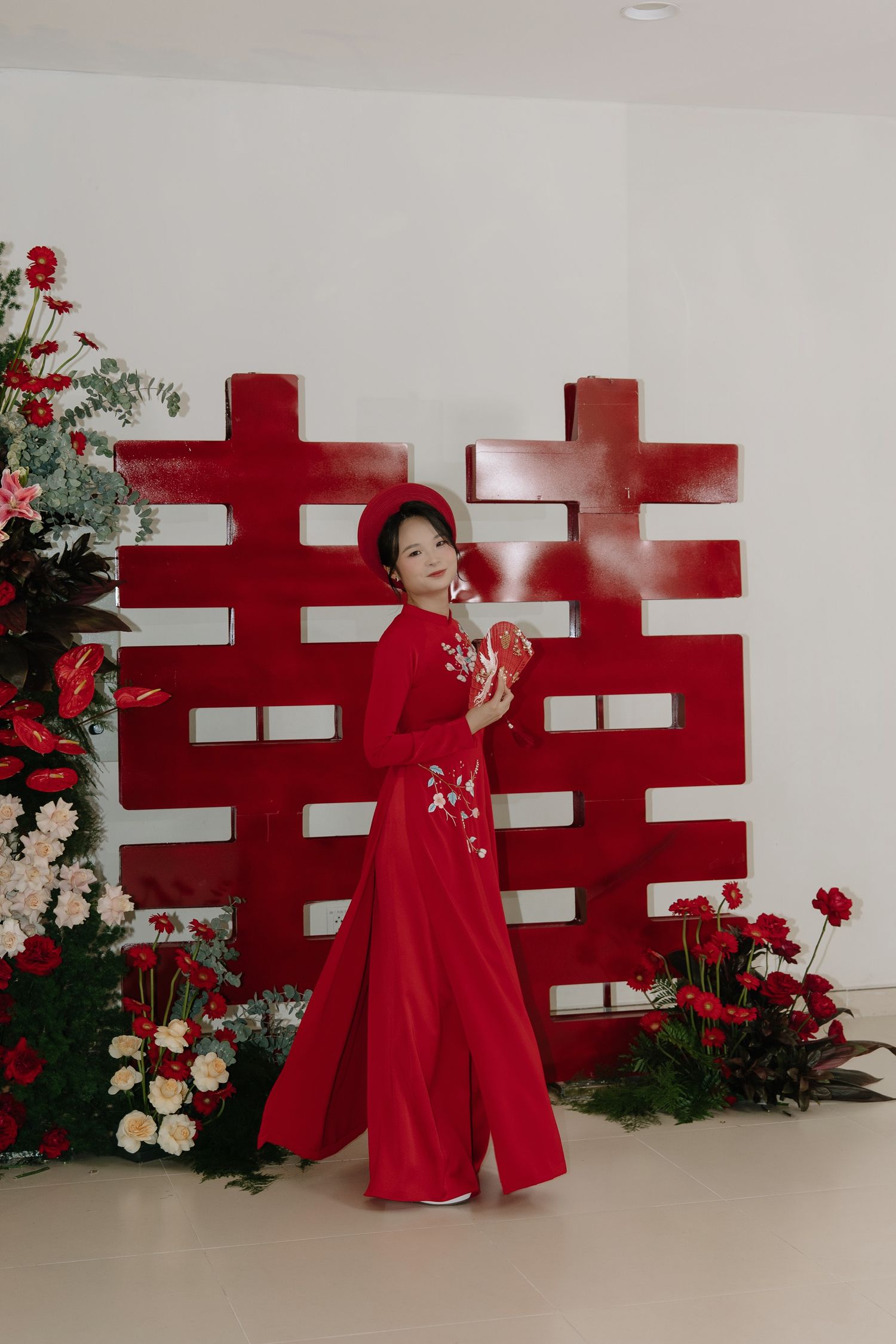 Bride in red ao dai gown poses before traditional double happiness symbol with floral arrangements.