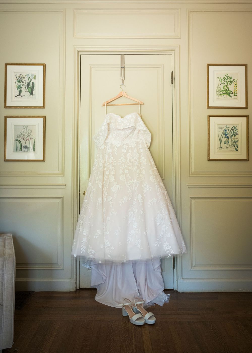 Wedding gown display showing a beautiful white dress hanging against cream paneled walls with framed artwork.