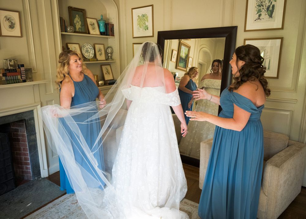 Bridesmaids in blue dresses help arrange the bride's veil in preparation room.