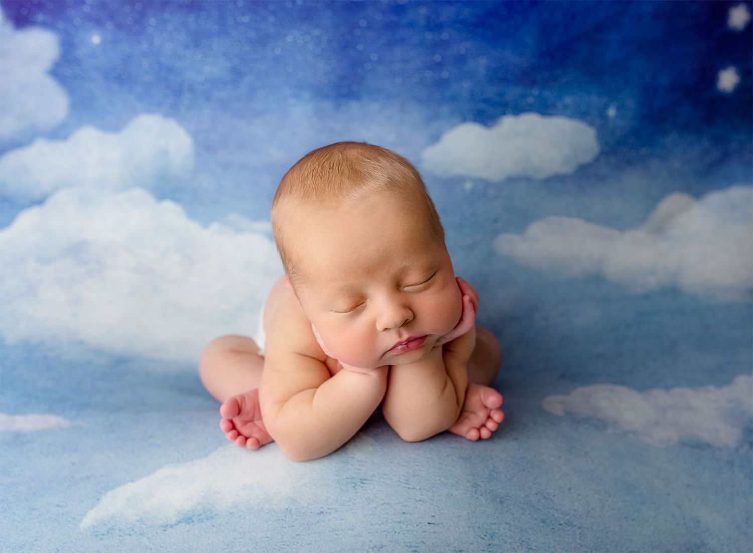A newborn baby sleeps peacefully on soft blue and white cloud-like fabric in a professional portrait setting.