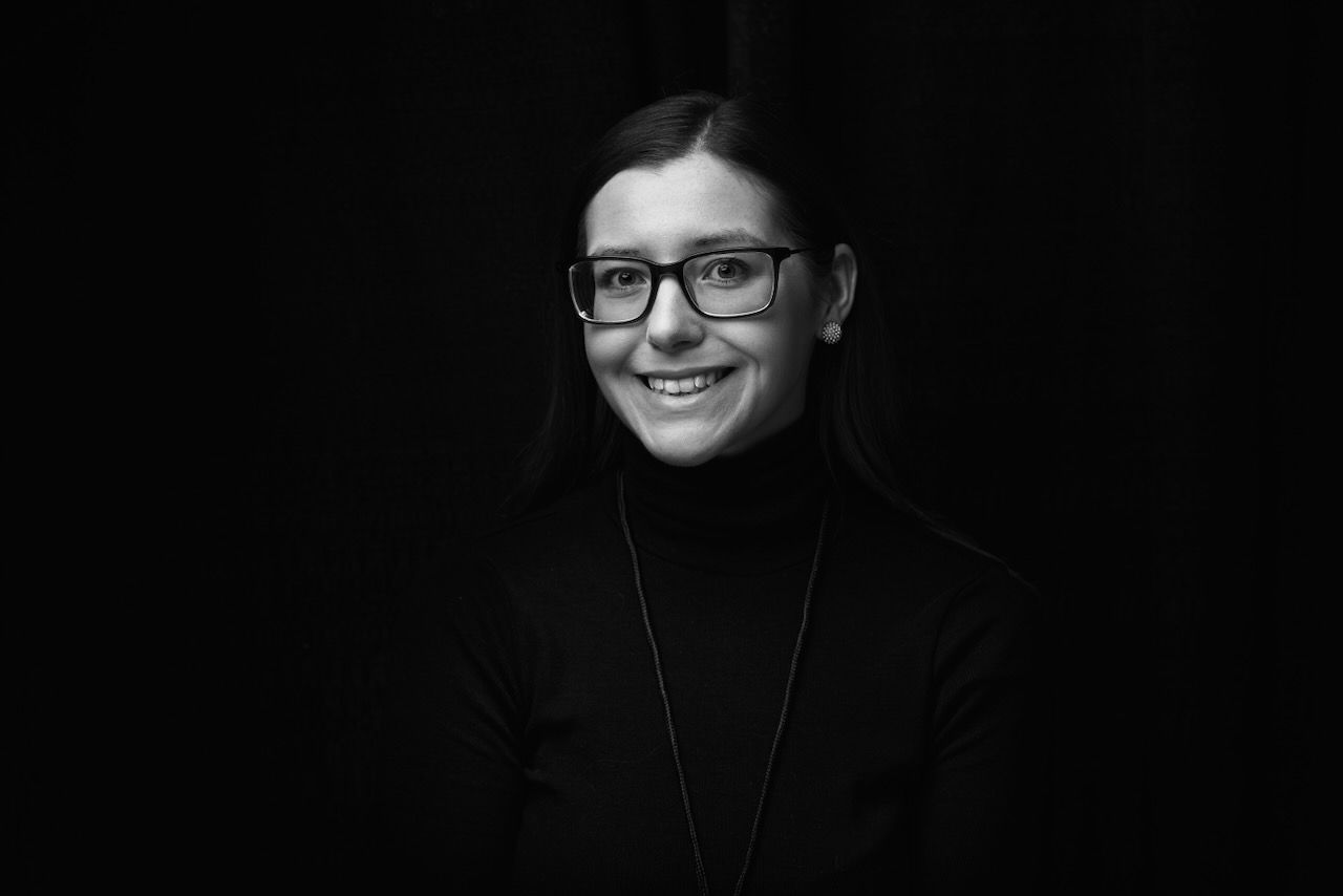 Black and white portrait of someone wearing glasses and a turtleneck sweater, smiling against dark background.