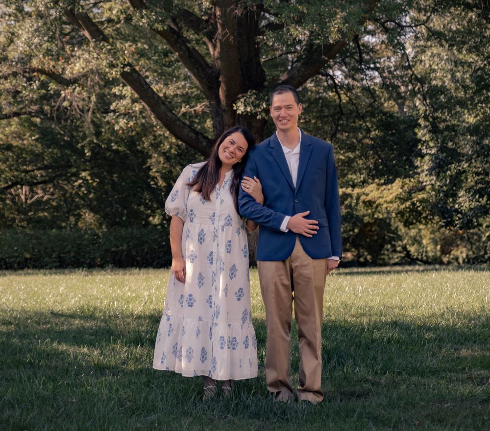 A couple stands together in a grassy field with trees in the background during a summer photo session.