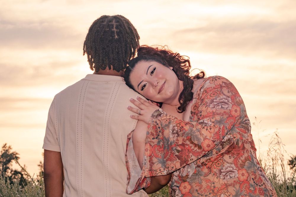 A couple embraces on a blanket in a field during sunset with warm orange sky in the background.