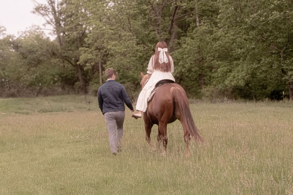 Couple walking together through a serene meadow with horses in the background.