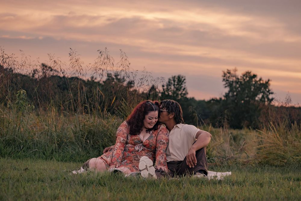 Two people share an intimate moment on a picnic blanket in a meadow at dusk with golden lighting.