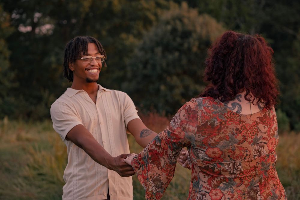 A couple holds hands and dances together in a golden field at sunset wearing white and floral outfits.