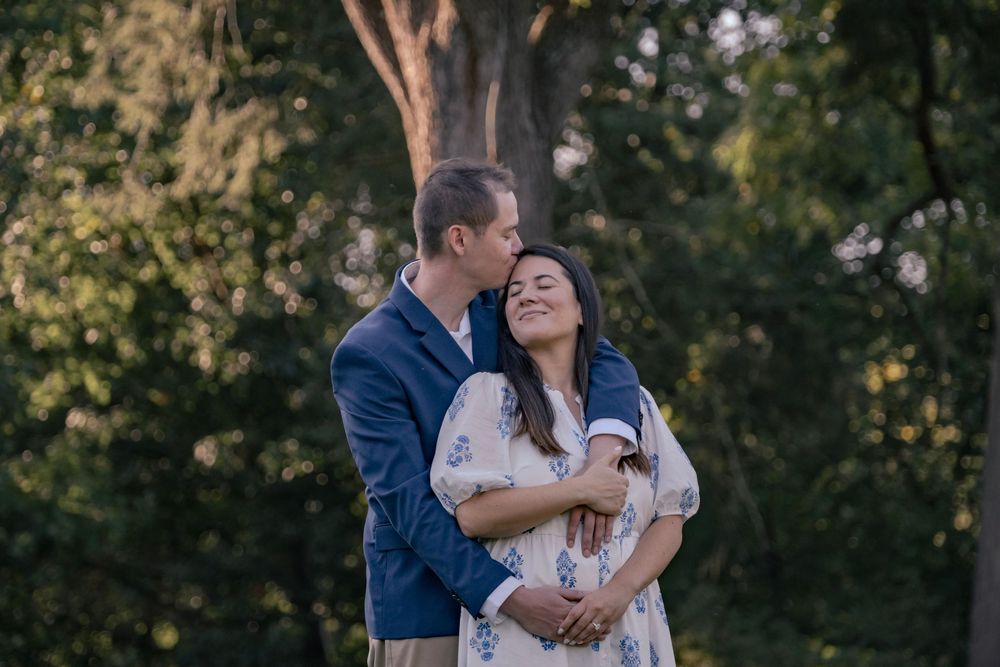 A couple embraces tenderly in a romantic outdoor setting surrounded by lush green trees during golden hour.