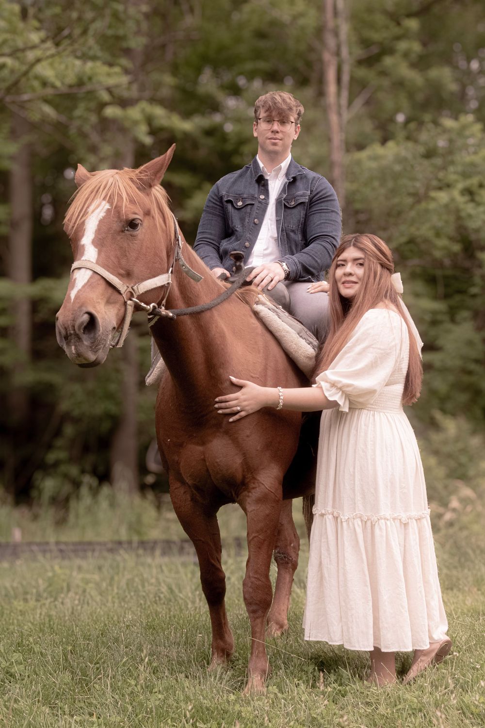 A couple poses with a brown horse in a grassy outdoor setting, with one person riding and one standing beside it.