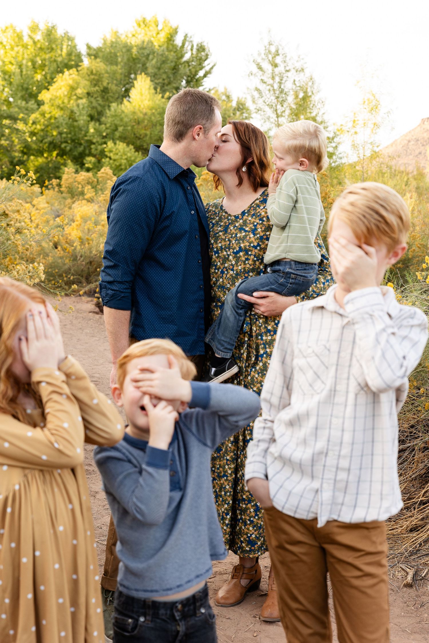 a mom and dat kissing with 4 kids blurry in the foreground covering their eyes. one small boy is also picking his nose