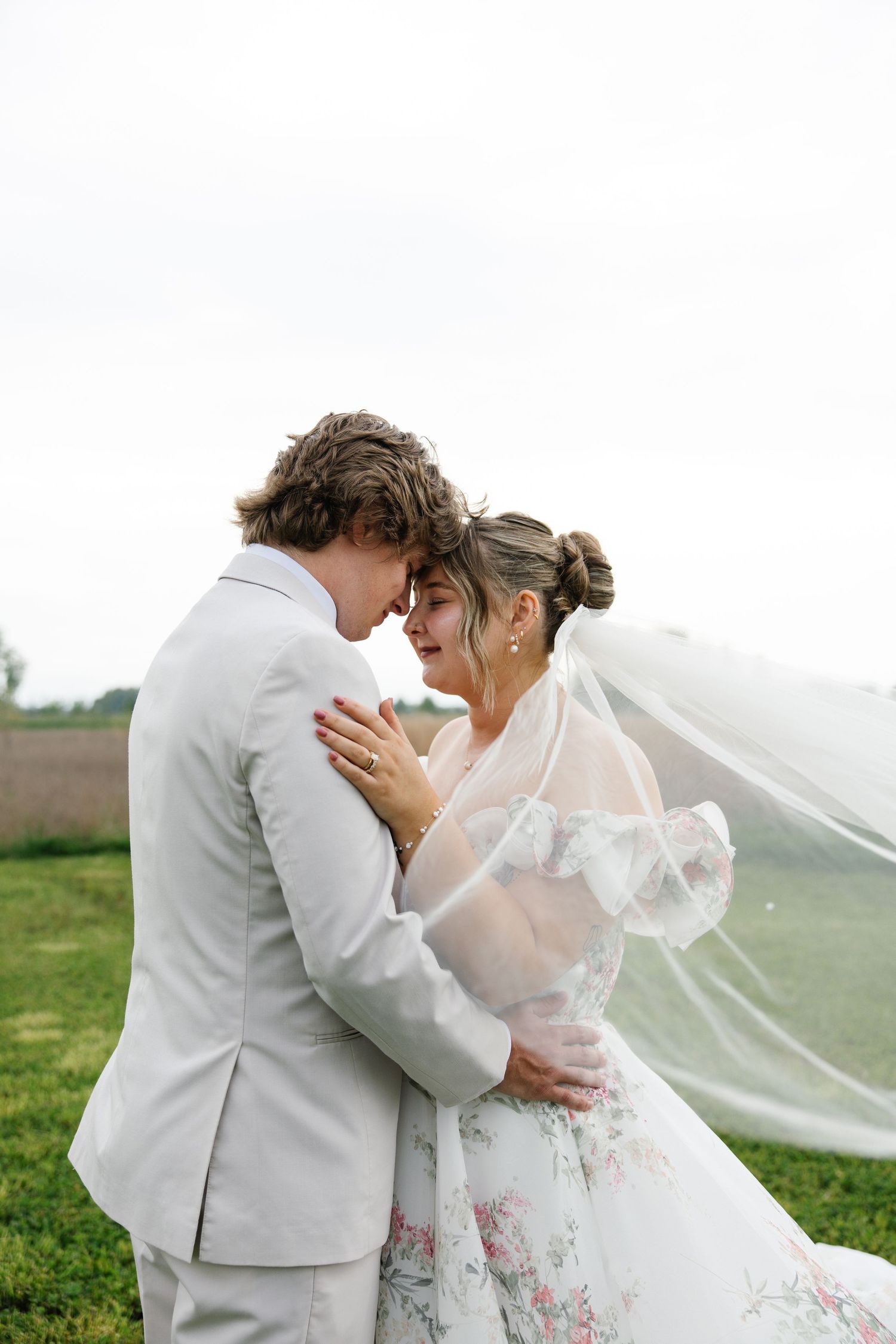 Wedding couple with clear umbrella and yellow bouquet embracing on grassy field.