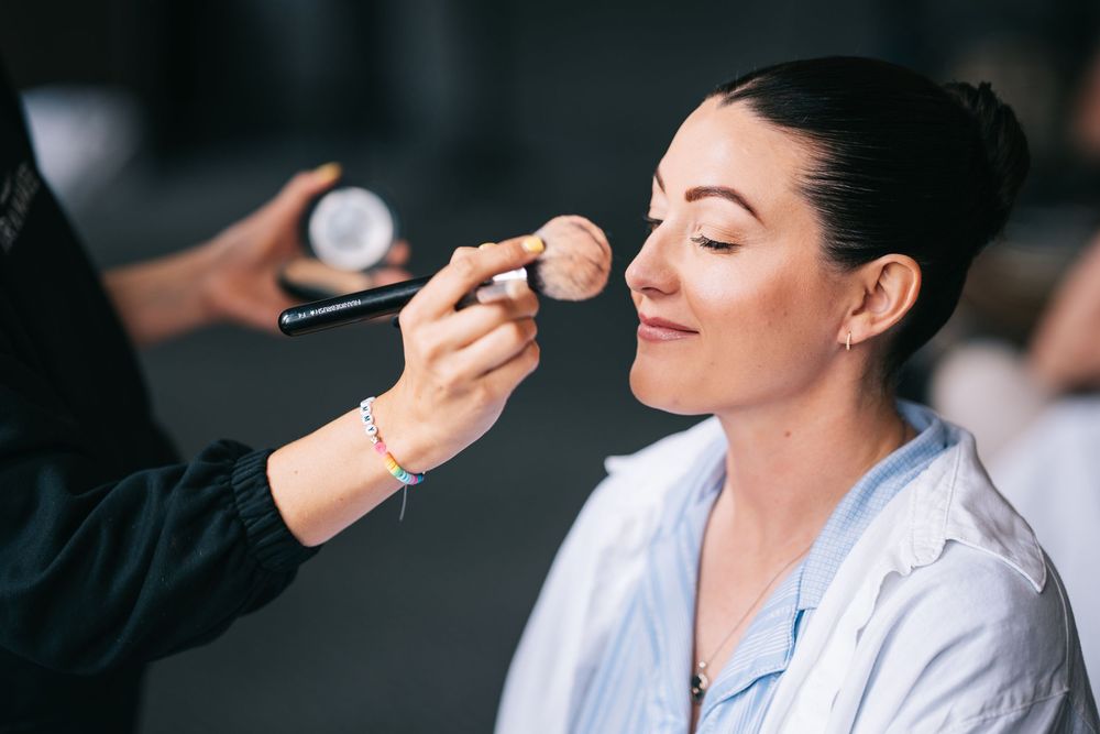A makeup artist applies cosmetics to a smiling client wearing a white shirt during a beauty session.