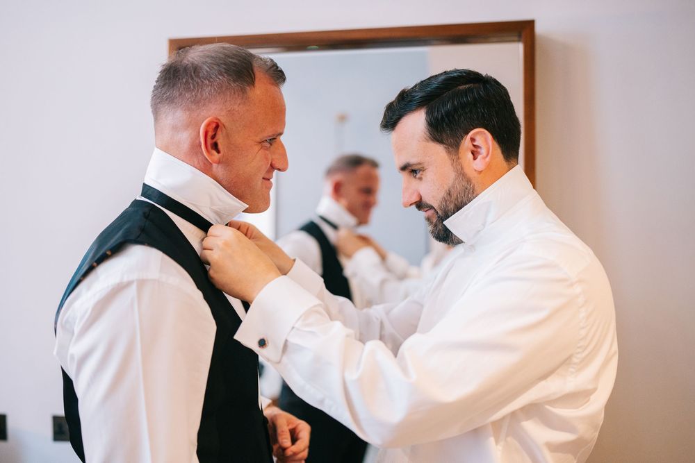 A groomsman helps adjust the white dress shirt of the groom while getting ready for a wedding ceremony.