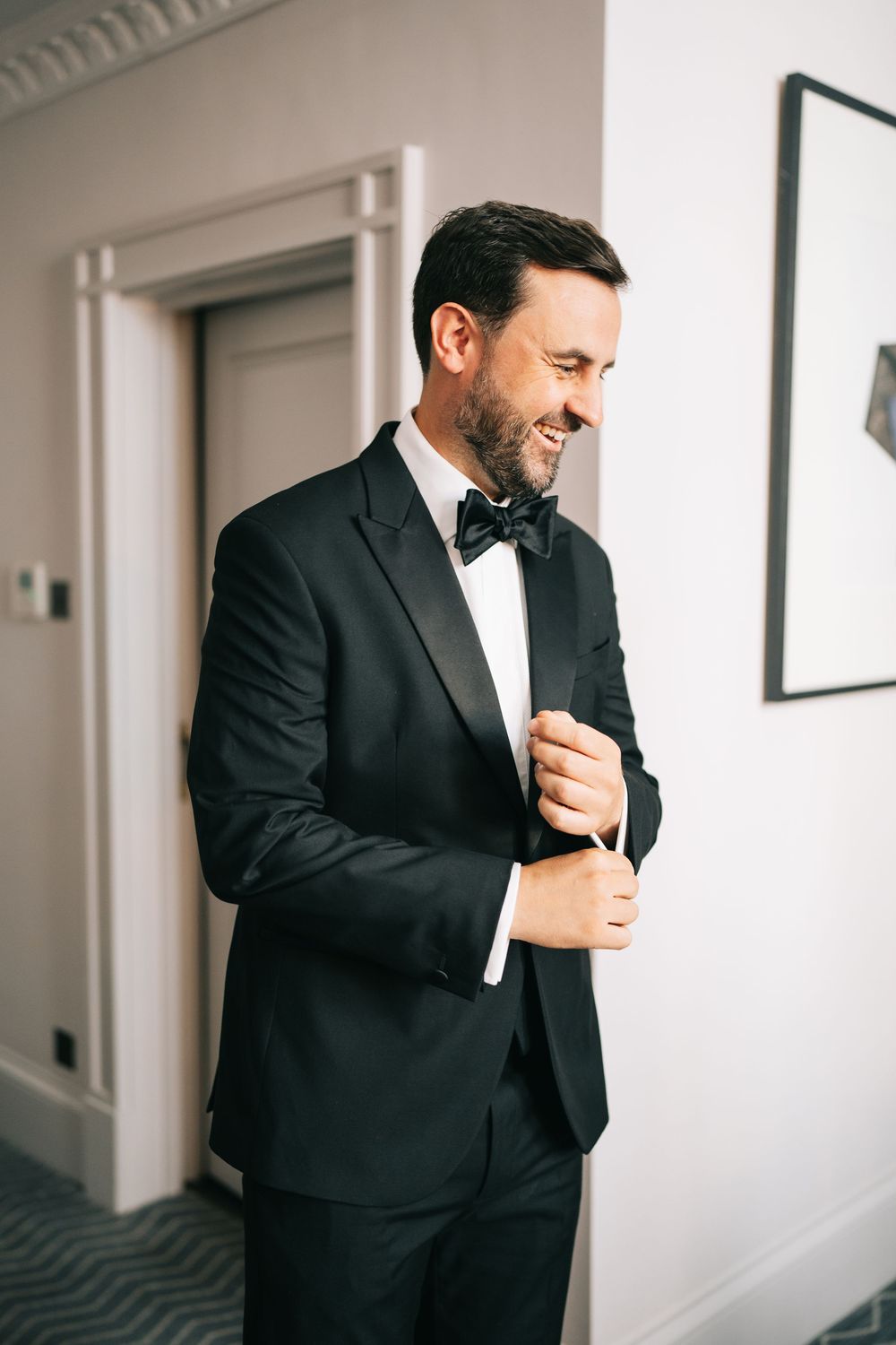 A person in a classic black tuxedo and bow tie smiles while adjusting their cuff in a hotel hallway.