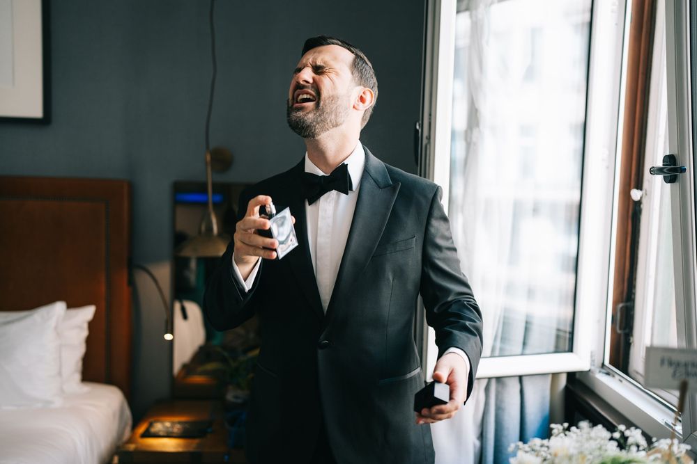 A person in a black formal suit jacket stands in a well-lit room next to a window.