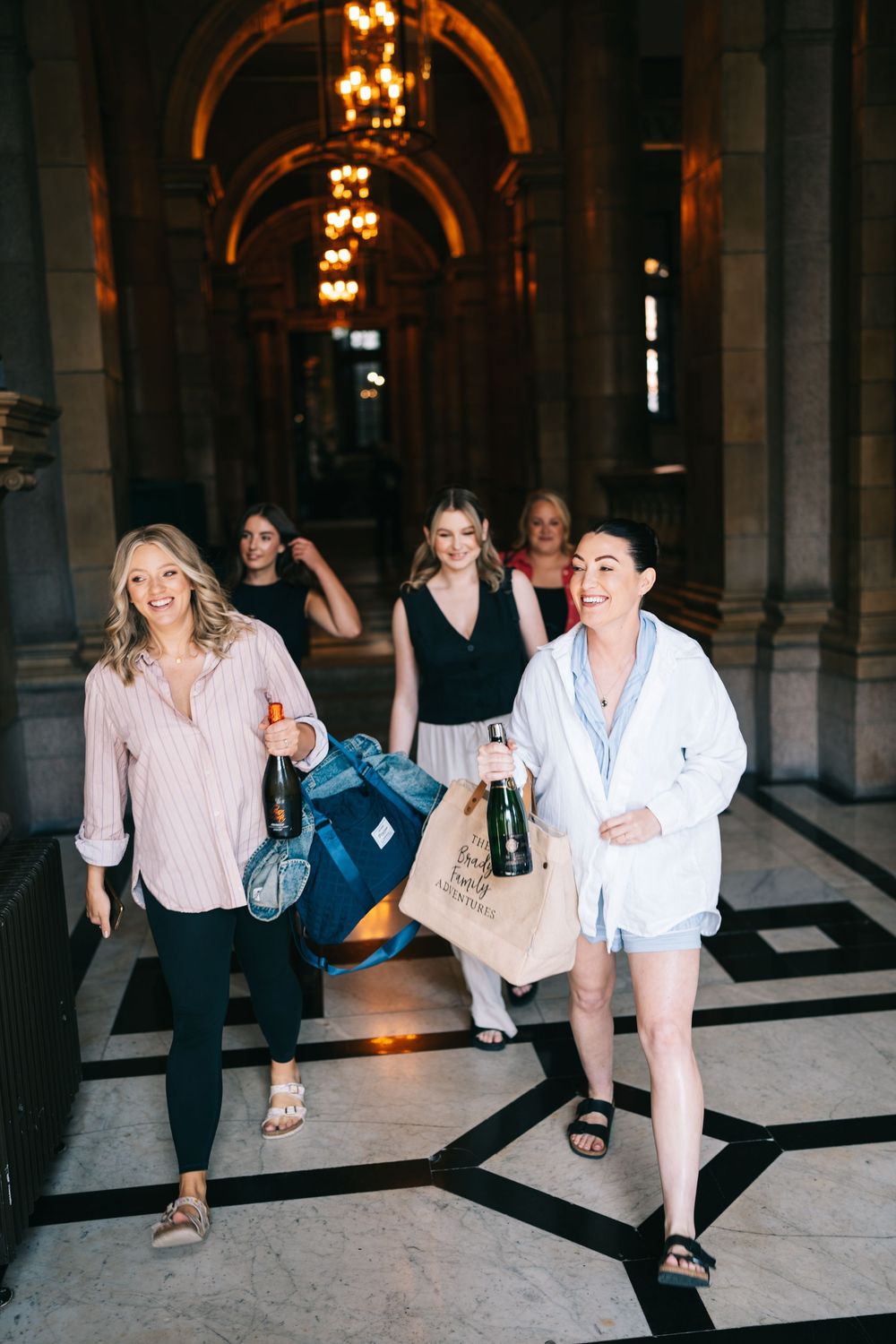 Group walking through an elegant hallway with arched ceilings and chandelier lighting carrying bags and drinks.