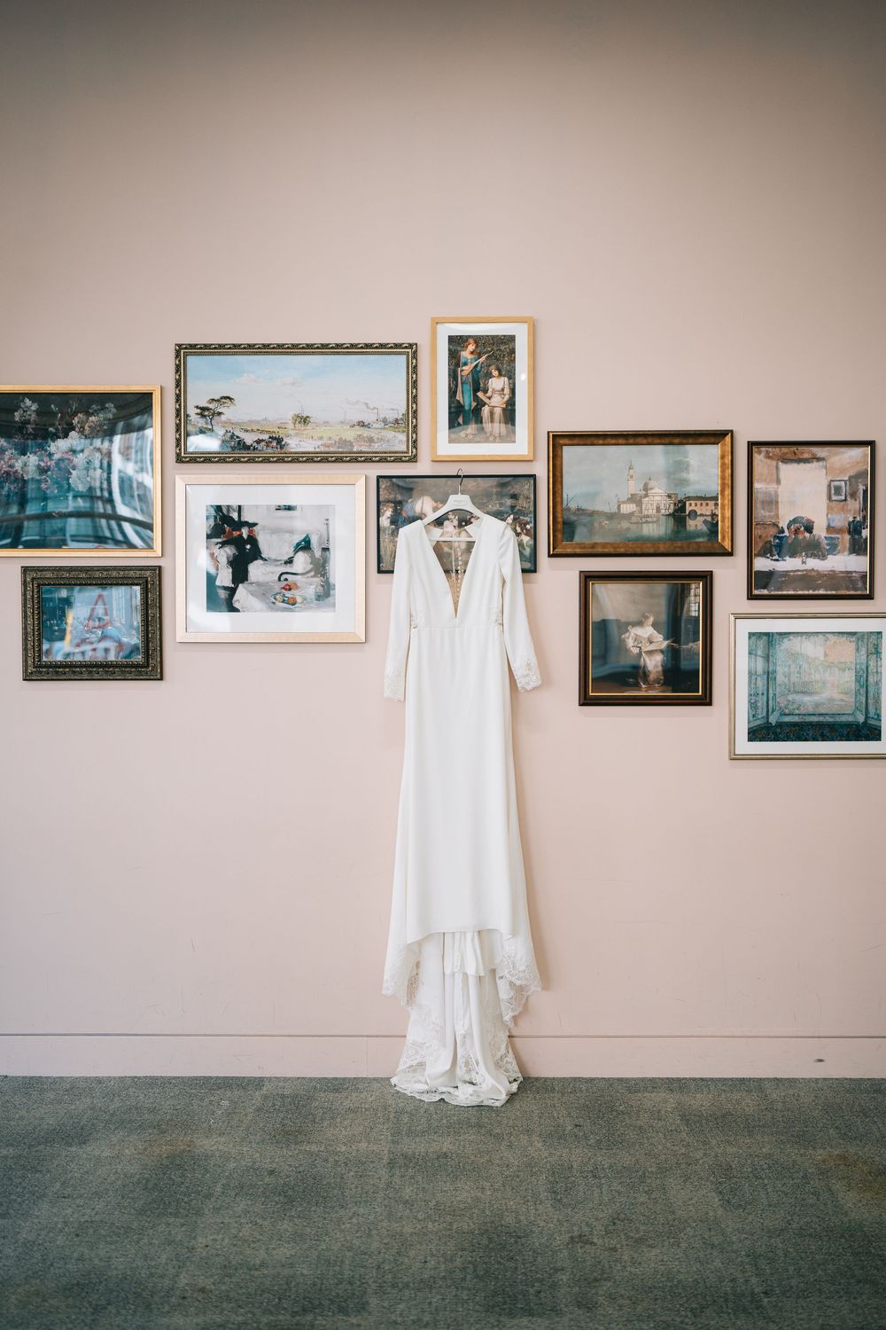 A white wedding dress hangs on a wall adorned with framed photographs in an art gallery setting.