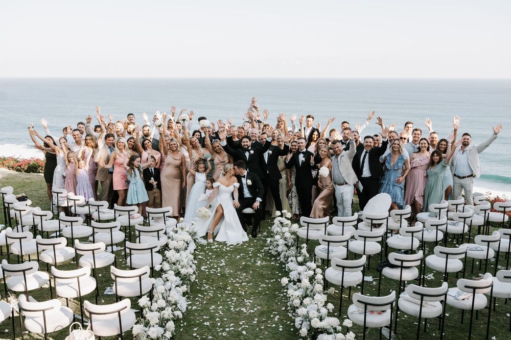 Group of wedding guests and bridal party jumping together on a beach in front of ocean at destination wedding celebration.