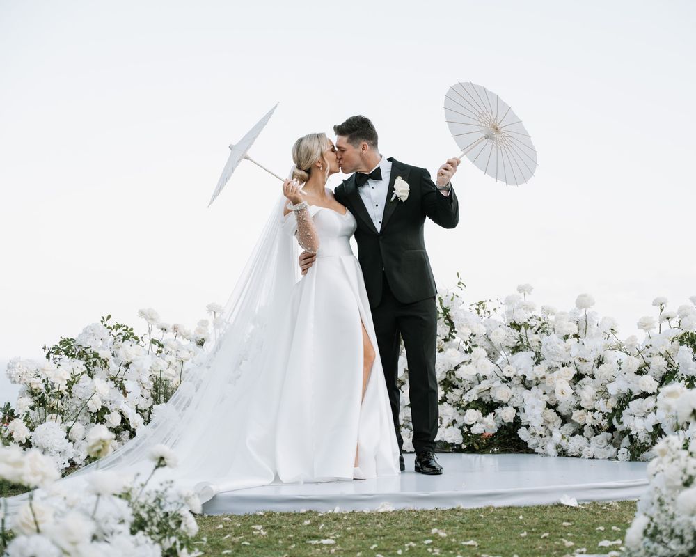 A romantic wedding couple holding white parasols pose together among white floral arrangements outdoors.