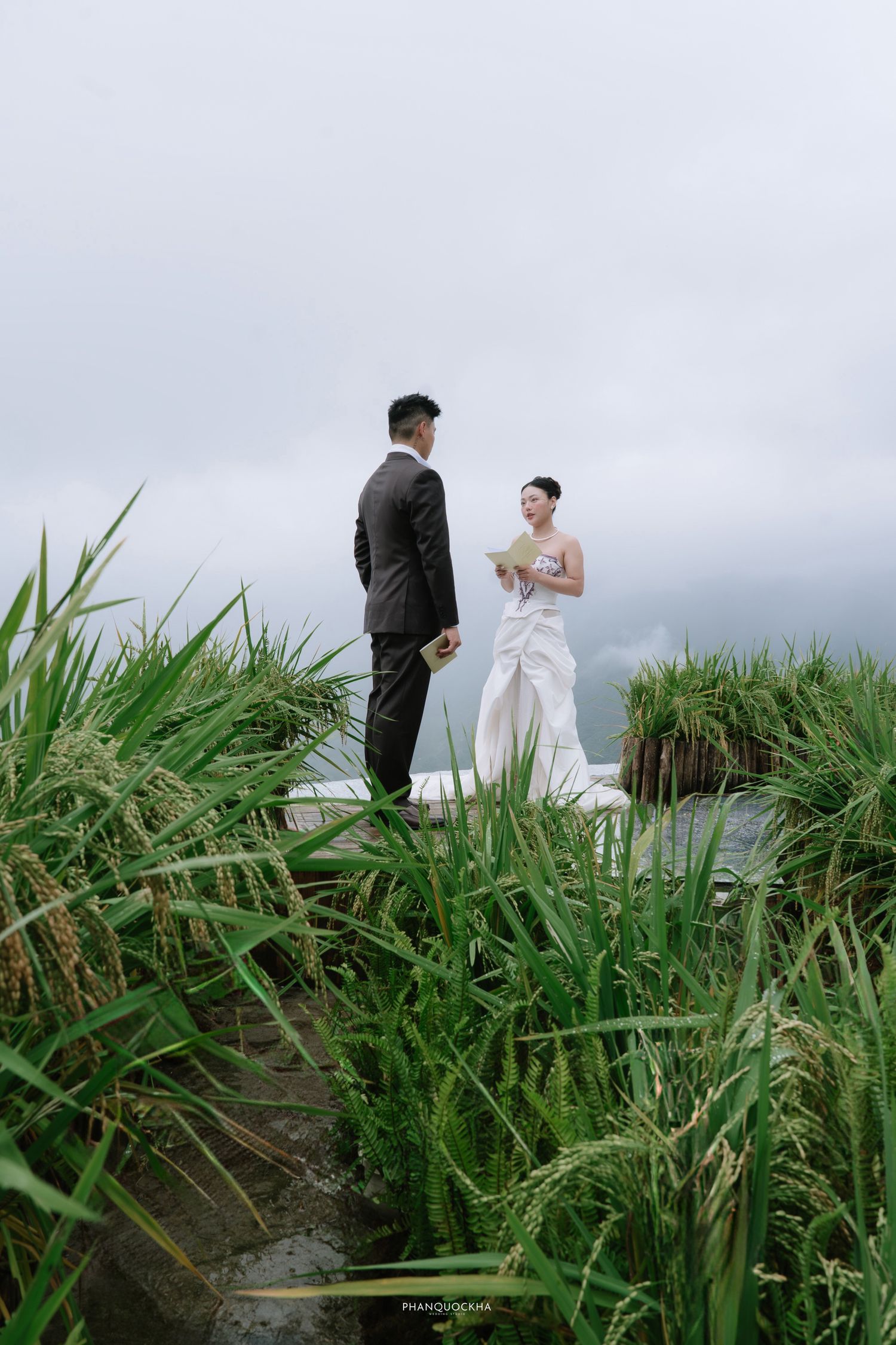 Bride and groom exchange vows in lush green rice field under overcast sky.