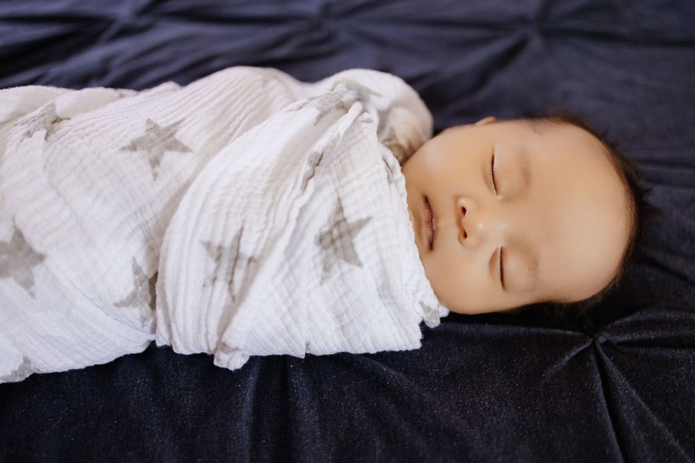 A newborn baby sleeps peacefully on a blue bedspread wrapped in a star swaddle during his in-home newborn photoshoot with Cindy Tucey