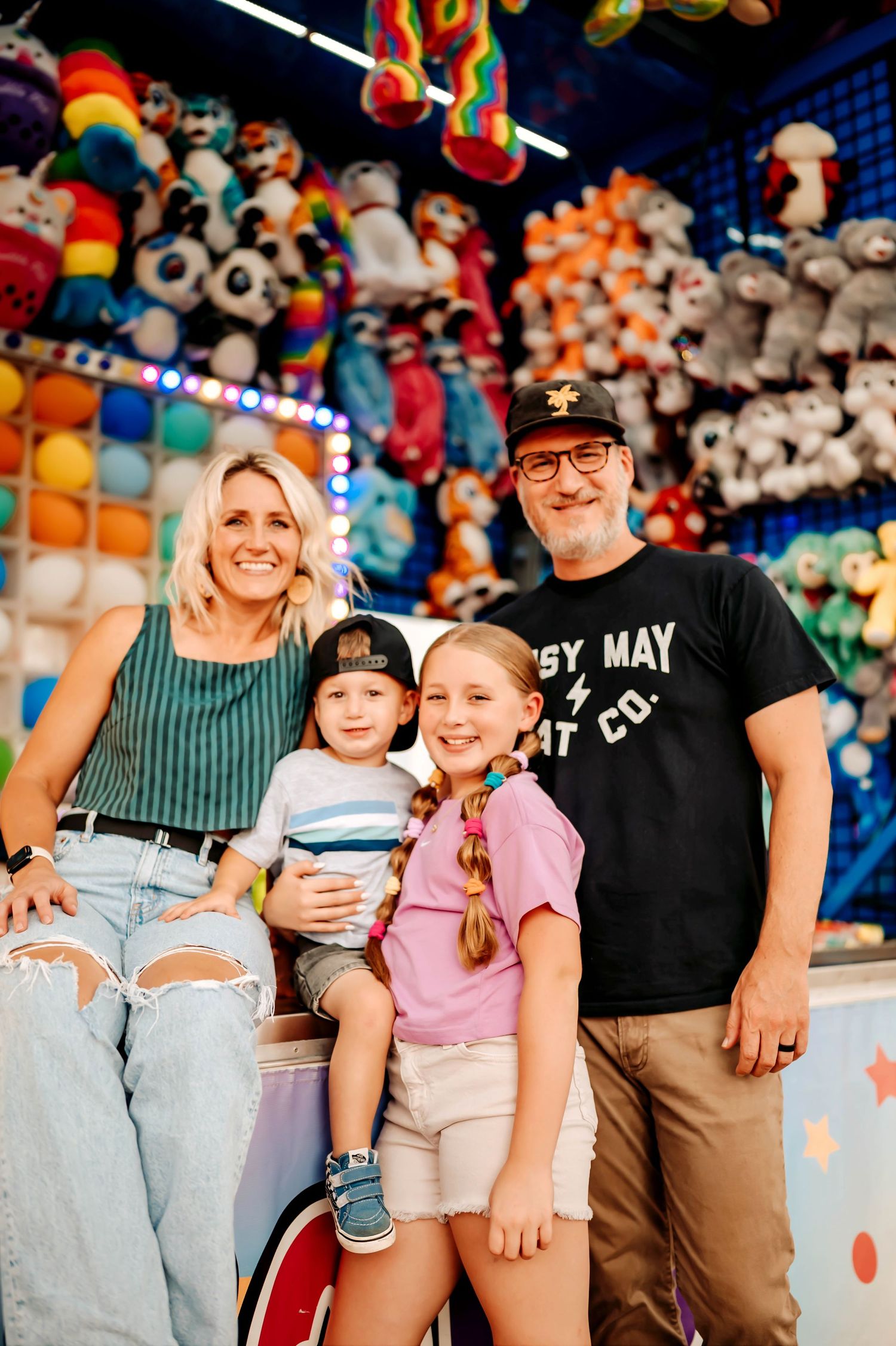 Family enjoying summer fun at carnival game booth with colorful stuffed animal prizes displayed in background.